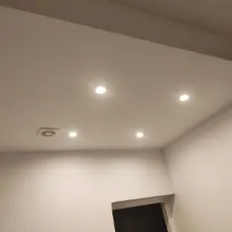 Białe wnętrze z wbudowanymi, punktowymi lampami LED w suficie i widocznym otworem wentylacyjnym, świeżo po malowaniu, z czarnym otworem drzwiowym w tle.