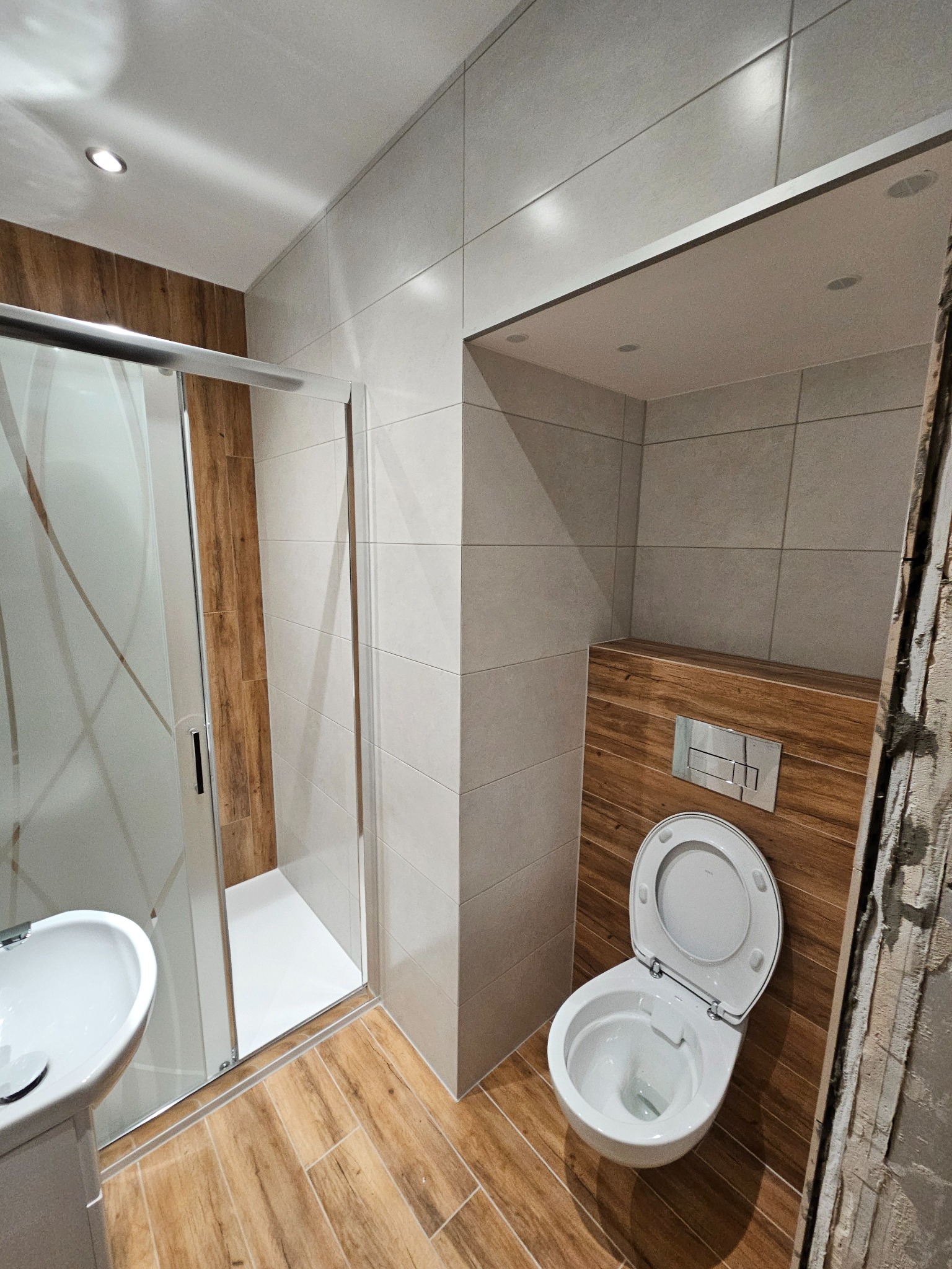 Nowoczesna łazienka po remoncie w Lądku-Zdroju. Kabina prysznicowa ze szklanymi drzwiami, umywalka i podwieszana toaleta z drewnianym akcentem. Jasne płytki na ścianach i podłodze.