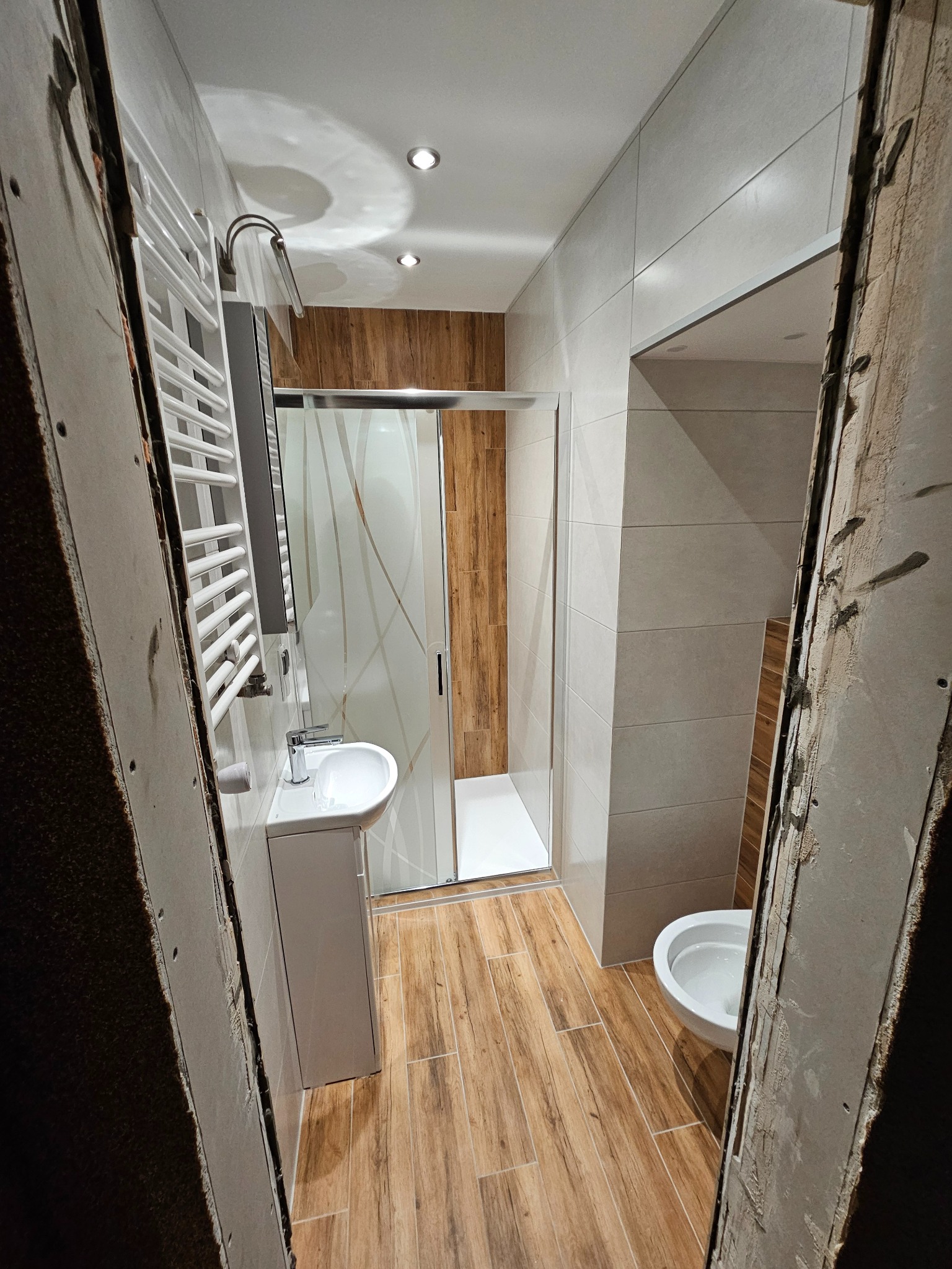 Wąskie ujęcie na wykończoną łazienkę z kabiną prysznicową, umywalką i grzejnikiem, widzianą przez surowy otwór w ścianie. Płytki imitujące drewno na podłodze i ścianach.