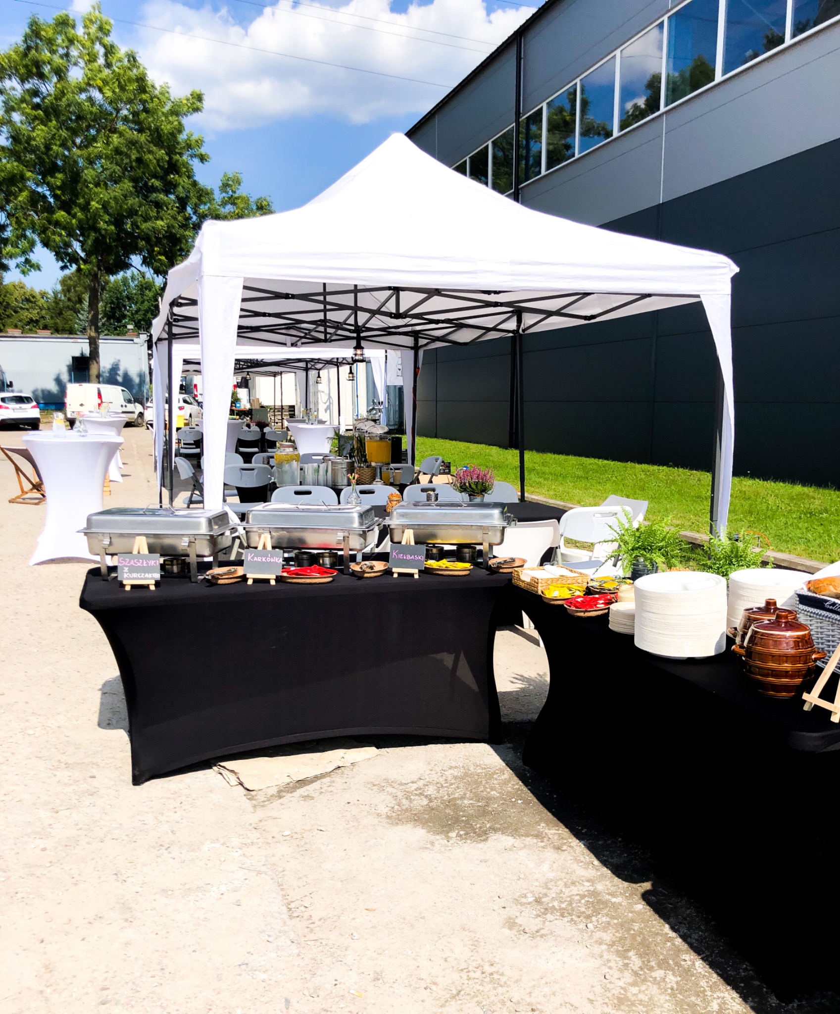 Zewnętrzny bufet z podgrzewaczami do potraw, napojami w szklanych słojach i dekoracjami kwiatowymi pod białym namiotem, z budynkiem w tle.