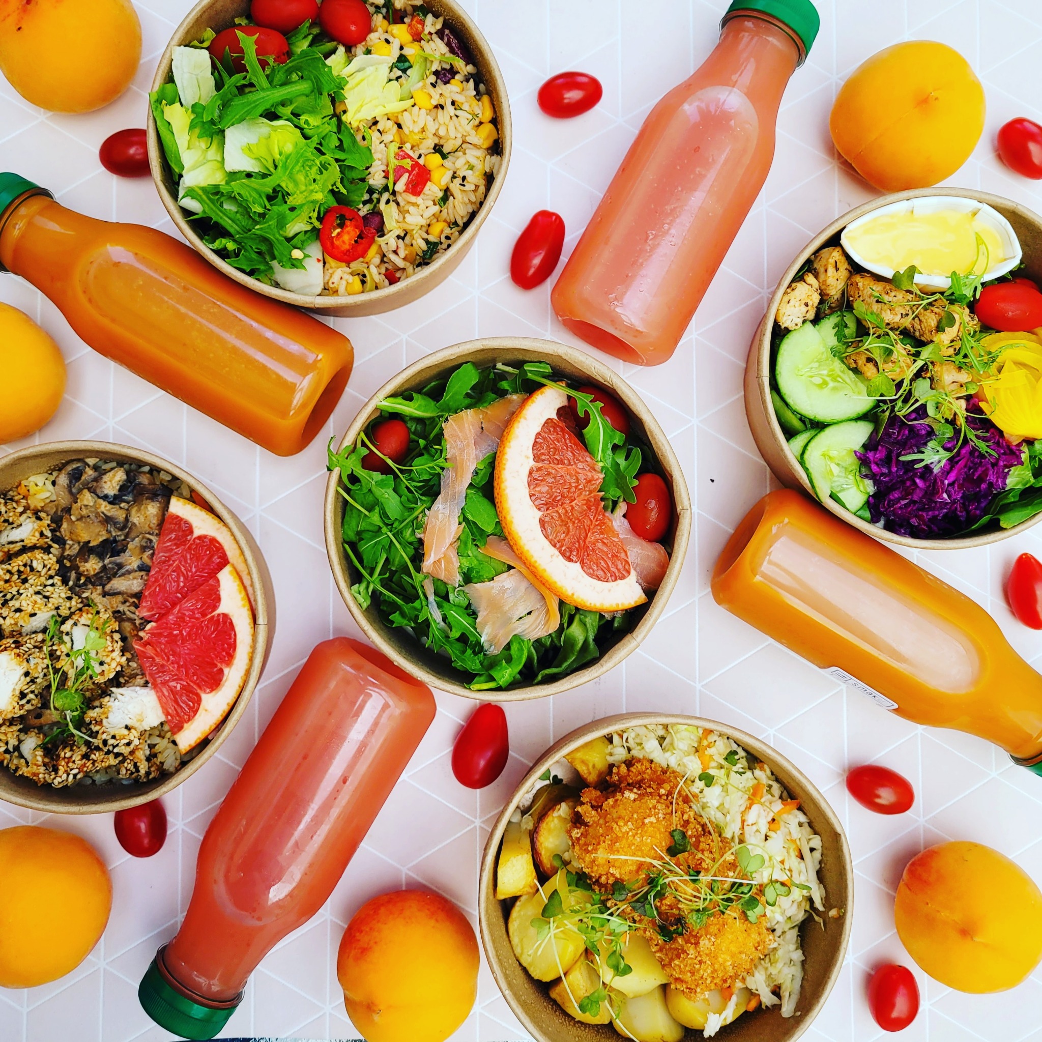 Zestaw pięciu misek z różnorodnymi sałatkami i dań oraz butelki ze świeżymi sokami na jasnym tle z geometrycznym wzorem, widok z góry.