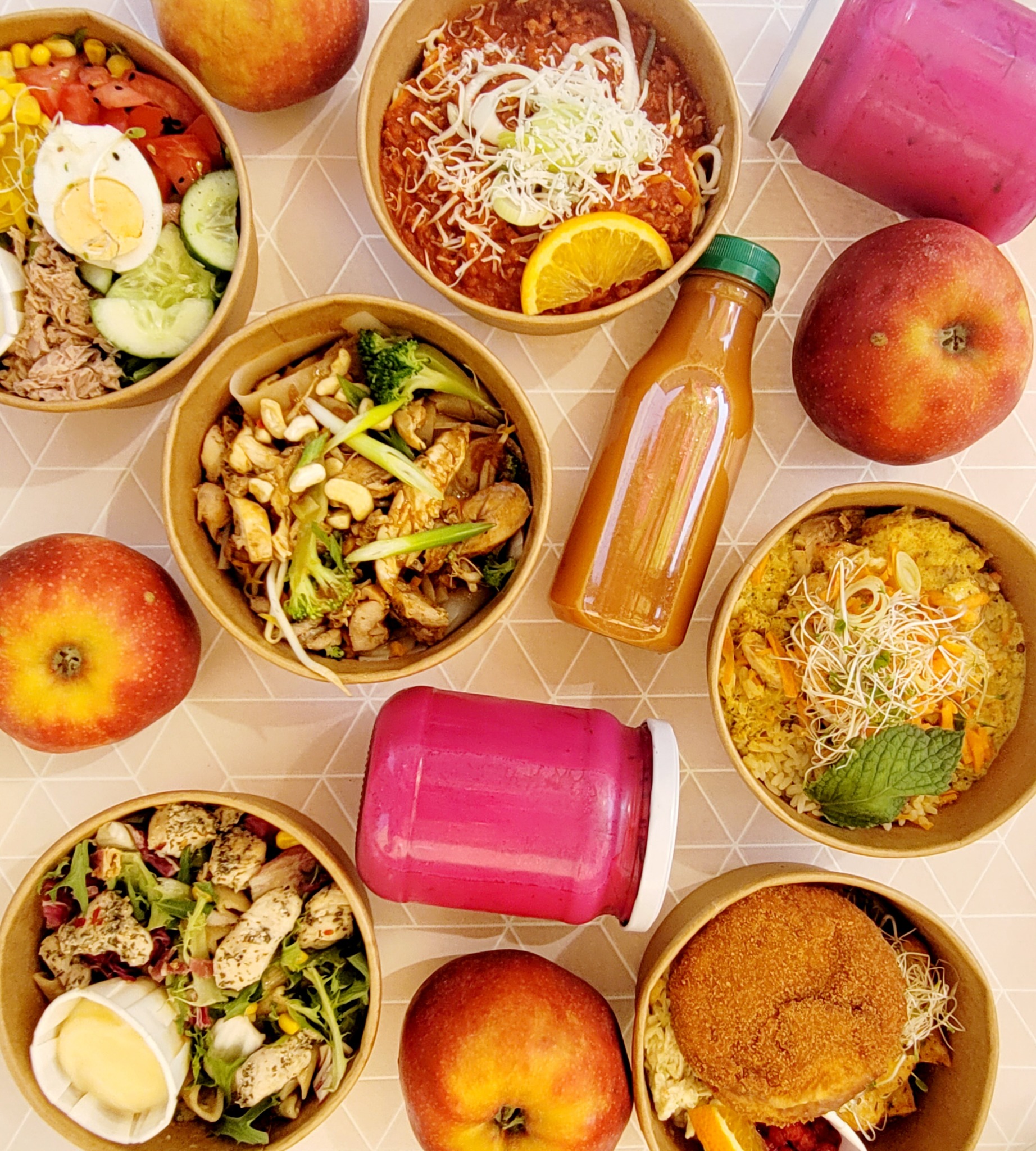 Różnorodne dania w papierowych miskach i butelkach z sokami, ułożone na geometrycznym tle, uzupełnione świeżymi jabłkami.