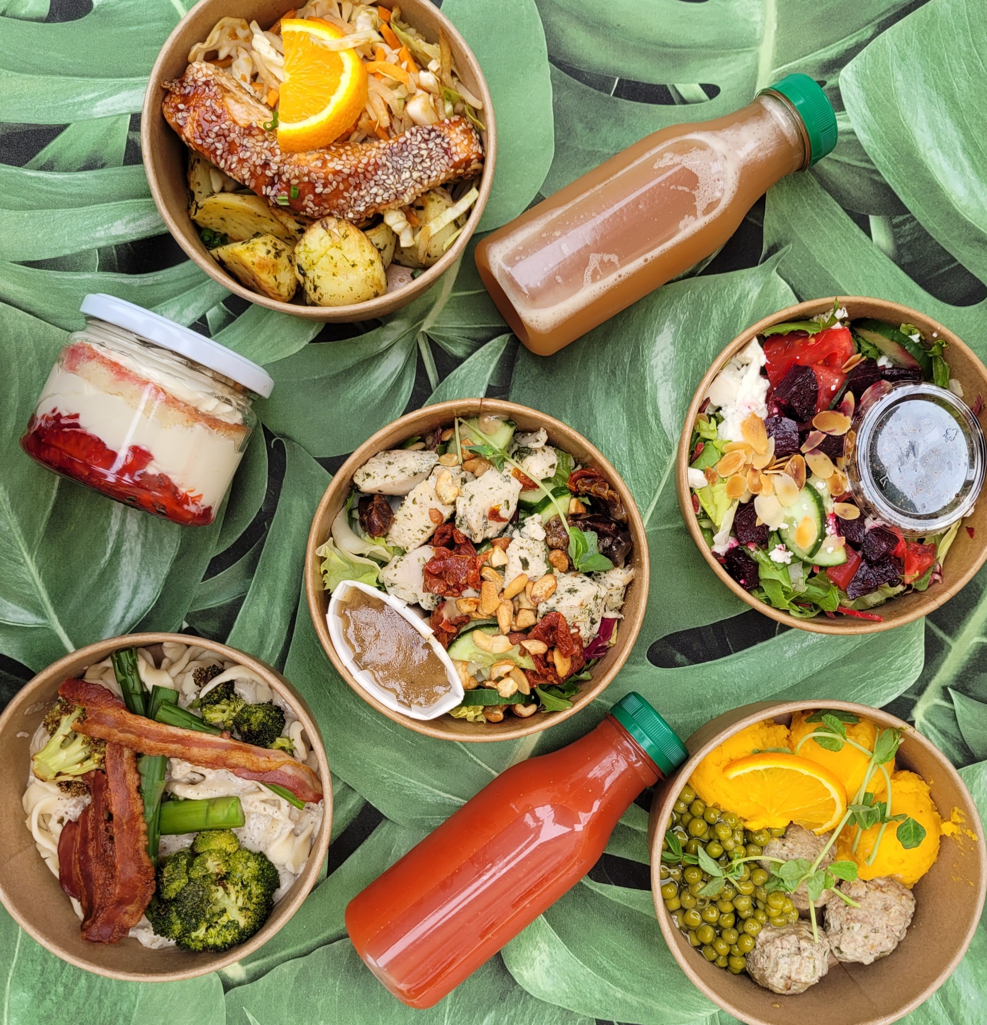 Zestaw sześciu dań w papierowych miskach i butelkach na tle liści: sałatki, danie z rybą w sezamie, deser.