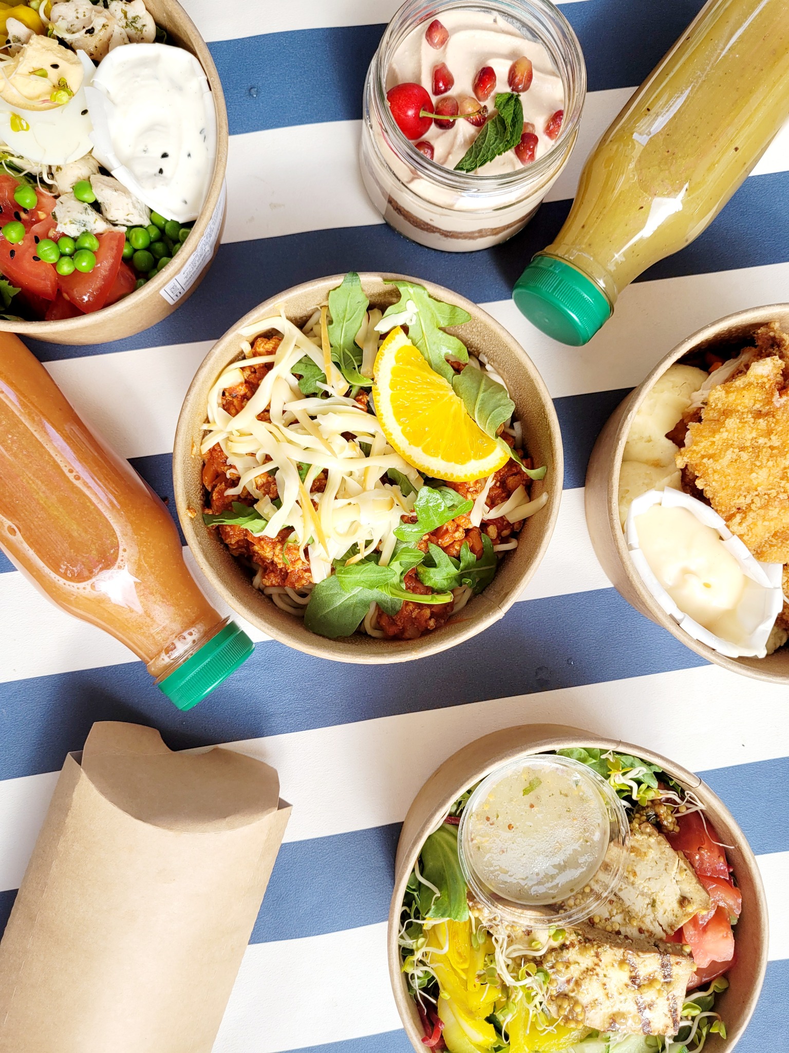 Zestaw pięciu dań w papierowych pojemnikach i dwóch butelek z sokiem na niebiesko-białym, pasiakowym tle. W pojemnikach widoczne różnorodne dania: sałatka z grillowanym tofu i warzywami, danie...