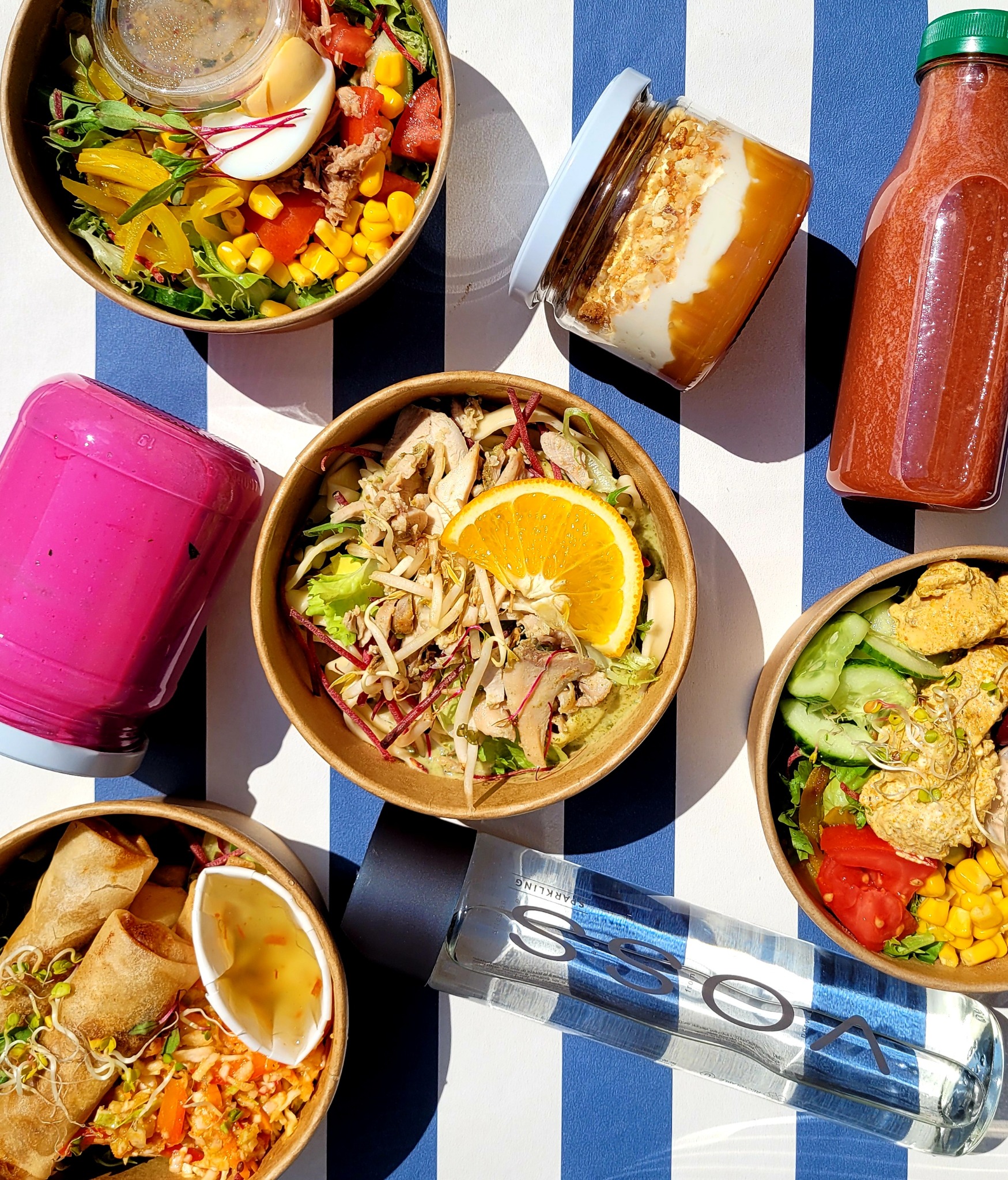 Zestaw różnorodnych dań w papierowych miskach i butelkach, obejmujący sałatki z warzywami i mięsem, spring rolls z sosem oraz napoje w butelkach na niebiesko-białym tle.