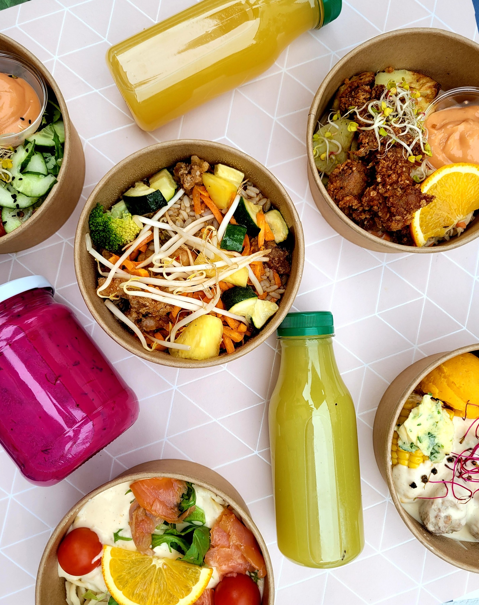 Kompozycja z sześciu dań w papierowych miskach i butelkach z sokami na różowym tle w geometryczne wzory. Dania zawierają różnorodne składniki, w tym warzywa, mięso, owoce i sosy.
