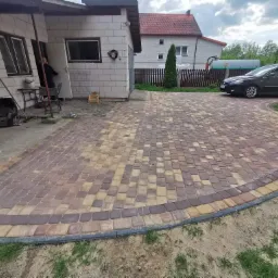 Nowo ułożony, półokrągły podjazd z kostki brukowej w odcieniach brązu i beżu, obrzeżony ciemniejszą kostką, prowadzący do garażu z białego bloczka; w tle dom z czerwonym dachem i zaparkowany czarny...