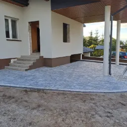 Nowoczesny podjazd z szarej kostki brukowej przed domem z kolumnami, widoczne schody wejściowe i fragment niezabudowanego terenu.