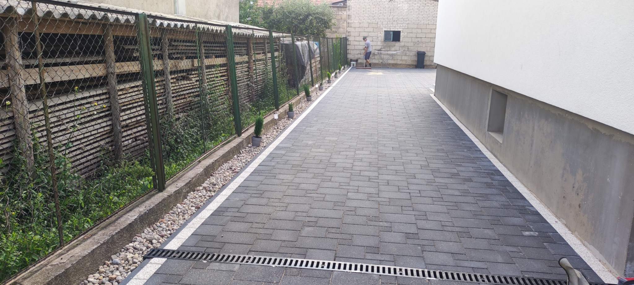 Ułożona szara kostka brukowa tworząca długi podjazd, oddzielona białym pasem od ściany budynku i ogrodzenia z siatki, z widocznym odwodnieniem liniowym i drobnymi roślinami wzdłuż granicy.