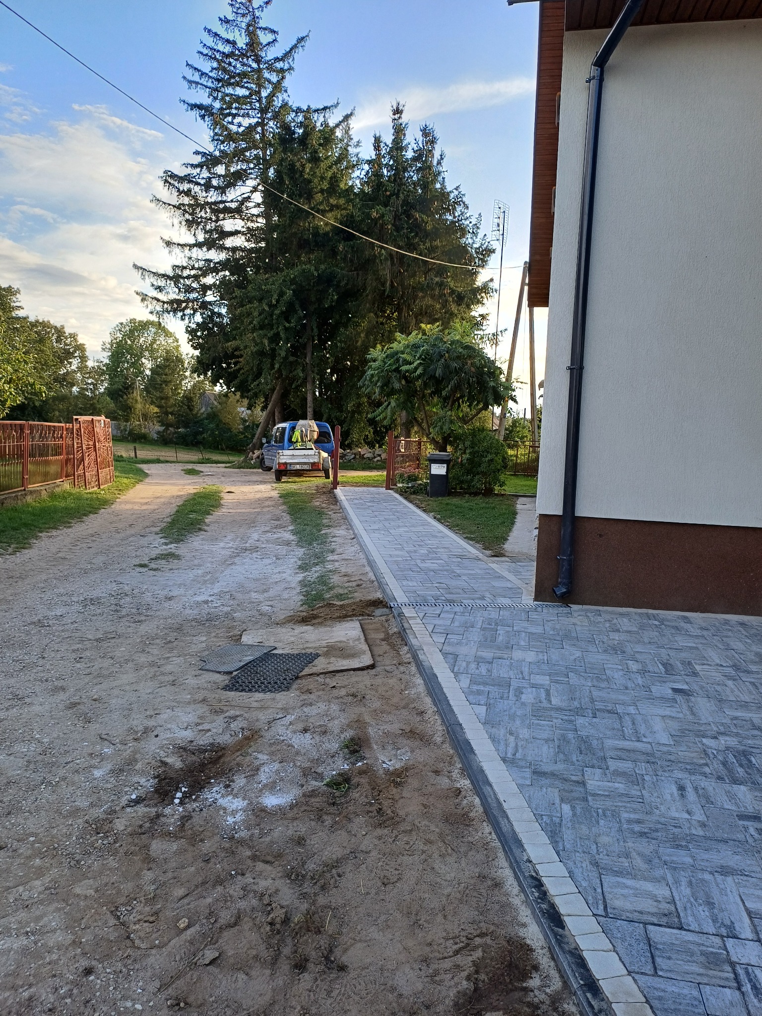 Ułożona szara kostka brukowa tworzy chodnik obok budynku. W tle widoczny samochód i drzewa. Częściowo wykonana usługa układania kostki.