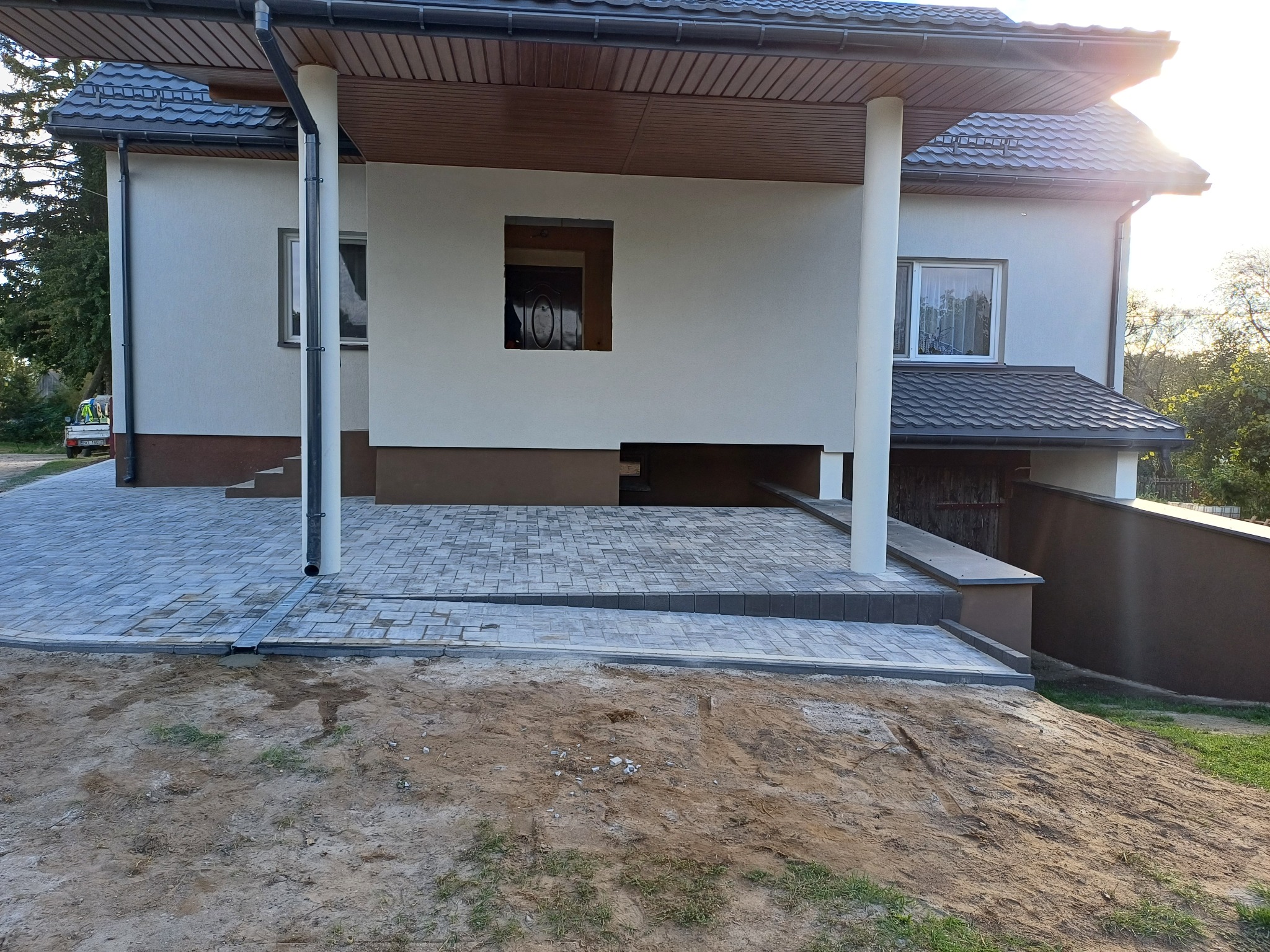 Nowoczesny podjazd z kostki brukowej w odcieniach szarości, z odpływem liniowym, prowadzący do domu z białymi kolumnami i brązowym wykończeniem.