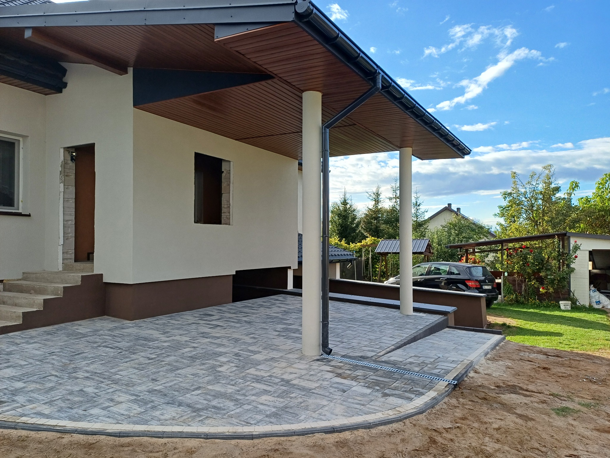 Nowoczesny taras z kostki brukowej w szarościach, z obrzeżami i odpływem liniowym, przylegający do domu z kolumnami podtrzymującymi zadaszenie.