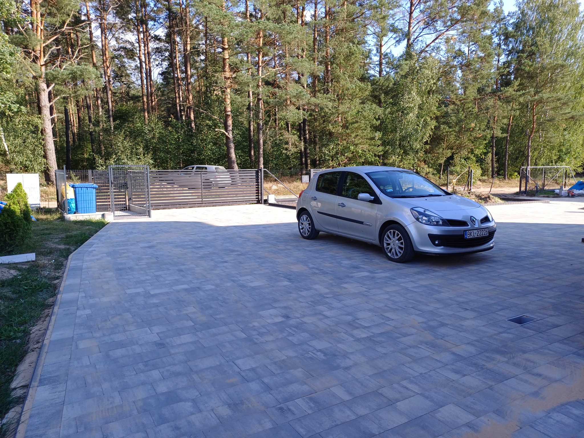 Nowoczesny podjazd z szarej kostki brukowej ułożonej we wzór prostokątny, z zaparkowanym srebrnym samochodem osobowym i bramą wjazdową w tle, otoczony zielenią drzew.