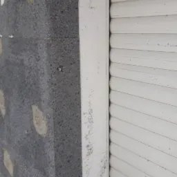 Fragment ściany z szarym styropianem elewacyjnym, widoczne kołki mocujące i biała rama okienna z roletą, na której widoczne są ślady zaprawy.