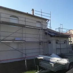 Dom jednorodzinny w trakcie tynkowania elewacji, widoczne rusztowania, przyczepka z materiałami budowlanymi i niezabezpieczony fragment dachu.