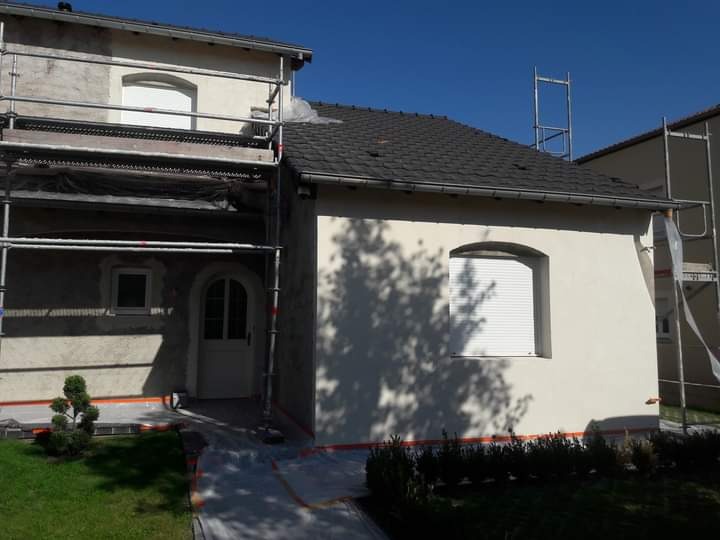 Dom jednorodzinny w trakcie prac elewacyjnych: część ściany otynkowana na biało, część surowa, rusztowania, zabezpieczenia folią, błękitne niebo.