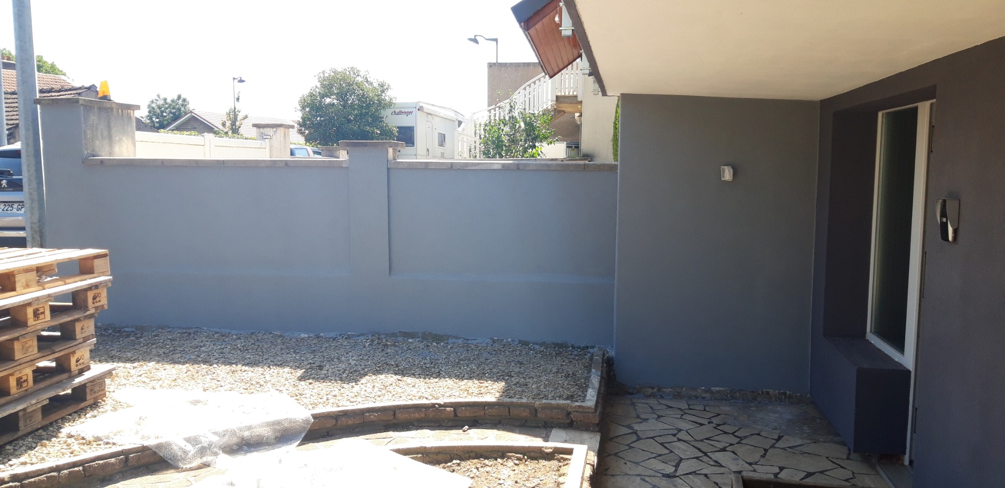 Świeżo pomalowany, gładki mur ogrodzeniowy w odcieniach szarości, z widocznymi paletami i żwirem u podstawy, przylegający do ściany budynku z oknem i kamienną ścieżką.