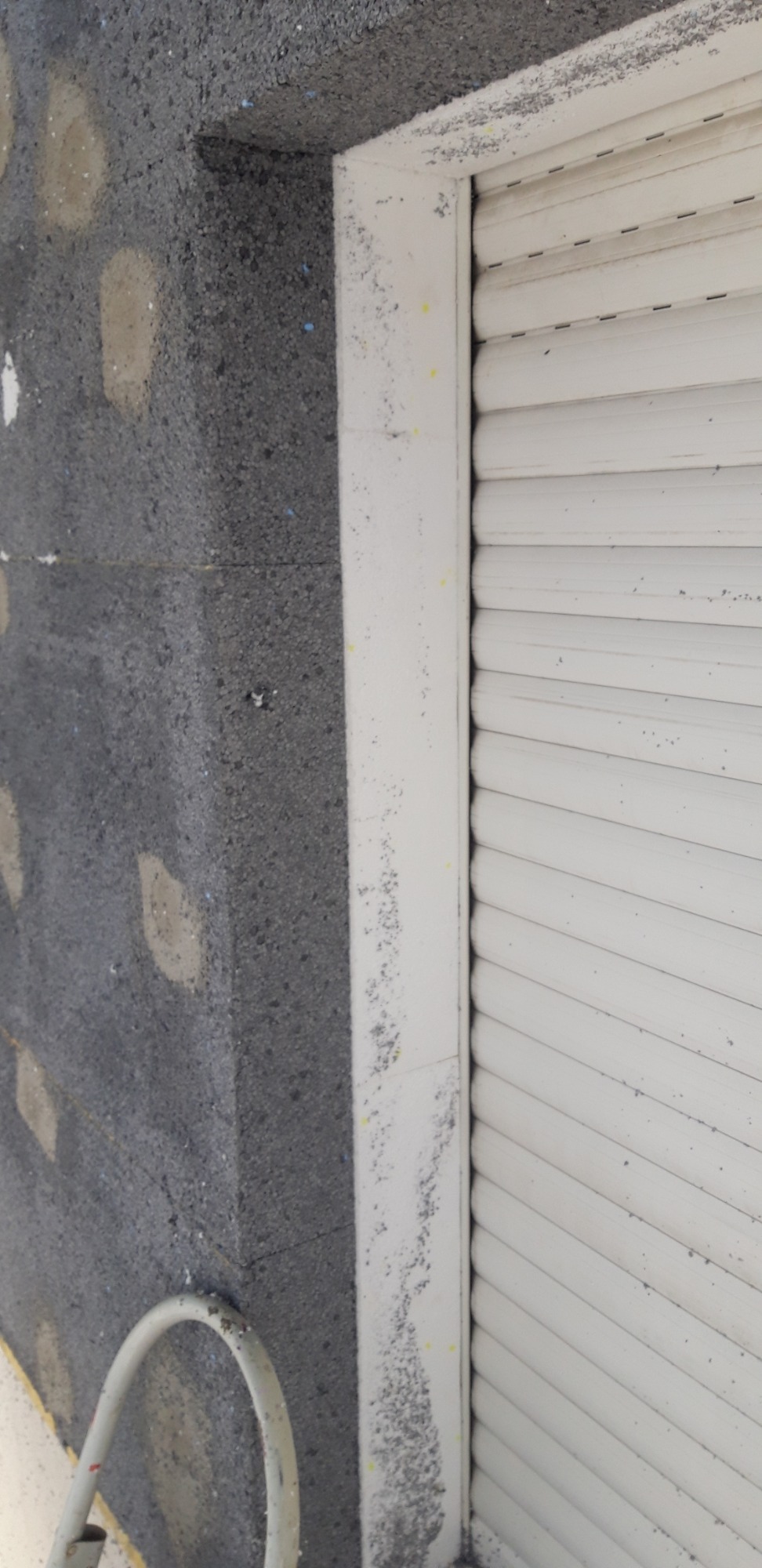 Fragment ściany z szarym styropianem elewacyjnym, widoczne kołki mocujące i biała rama okienna z roletą, na której widoczne są ślady zaprawy.
