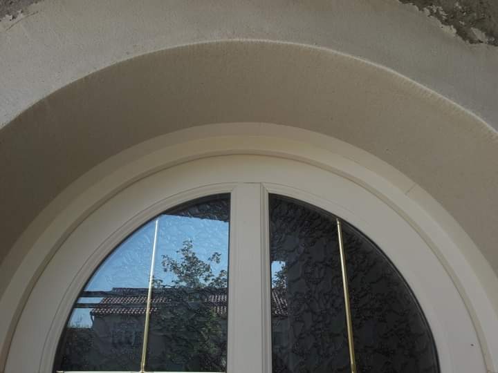 Fragment elewacji z łukowym oknem, widoczna struktura tynku i odbicie drzewa w szybie.