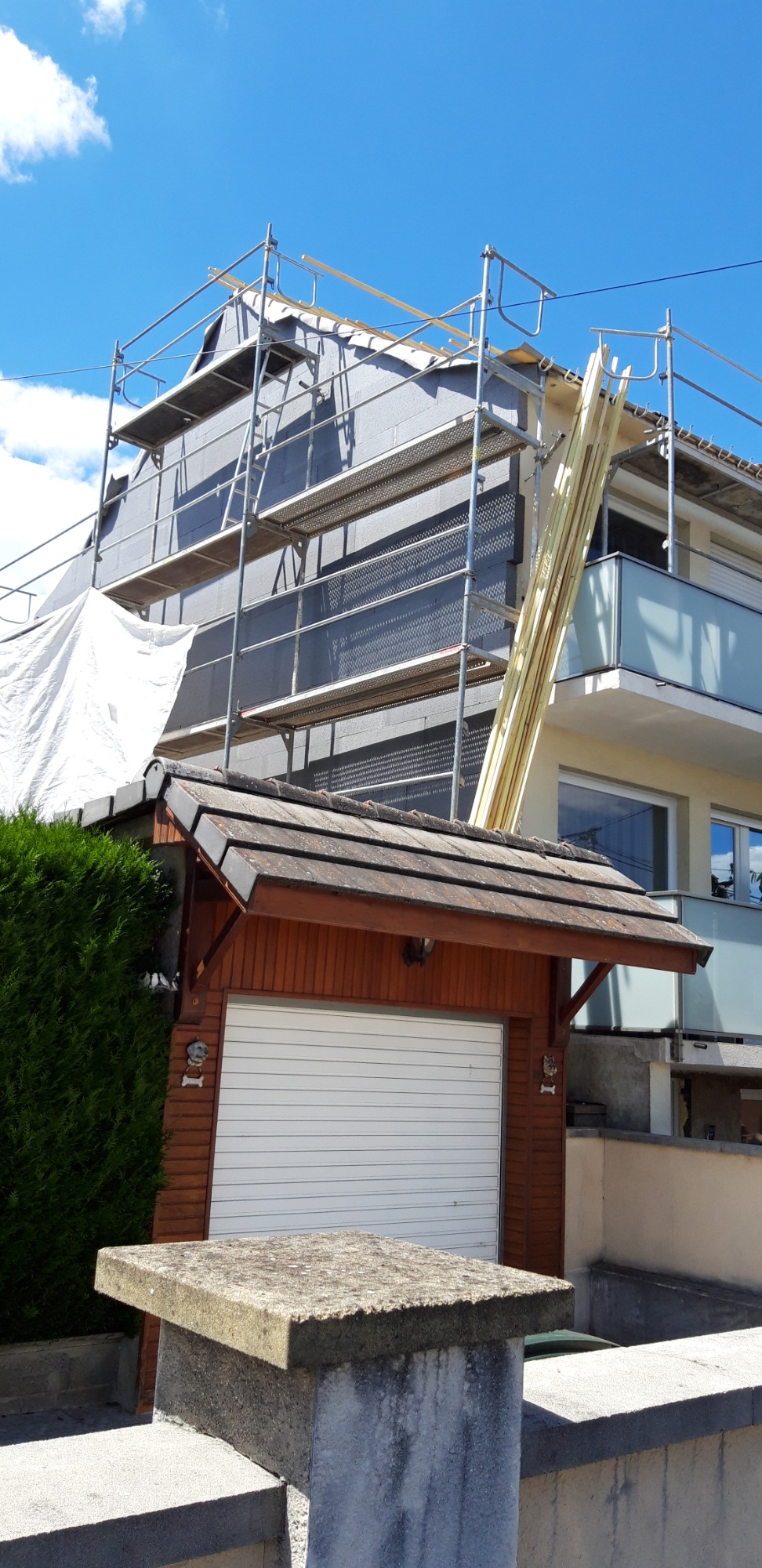 Dom jednorodzinny w trakcie ocieplania, widoczne rusztowania, styropianowe płyty elewacyjne i deski opierające się o rusztowanie, garaż z dekoracyjnym elementem w kształcie głowy psa.