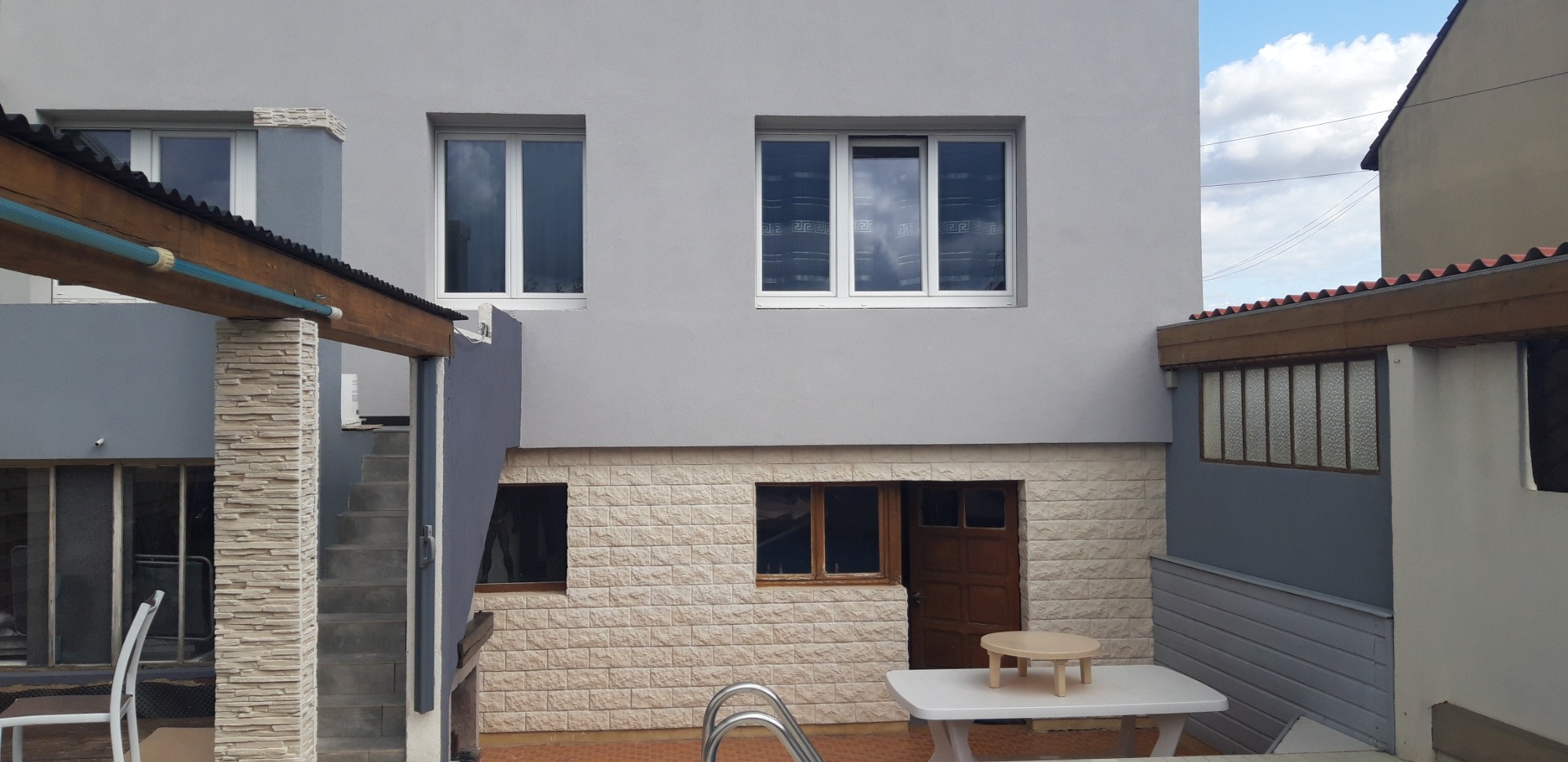 Szary budynek z oknami i drzwiami, wykończony kamieniem dekoracyjnym na dole elewacji, widok na fragment podwórka z basenem.