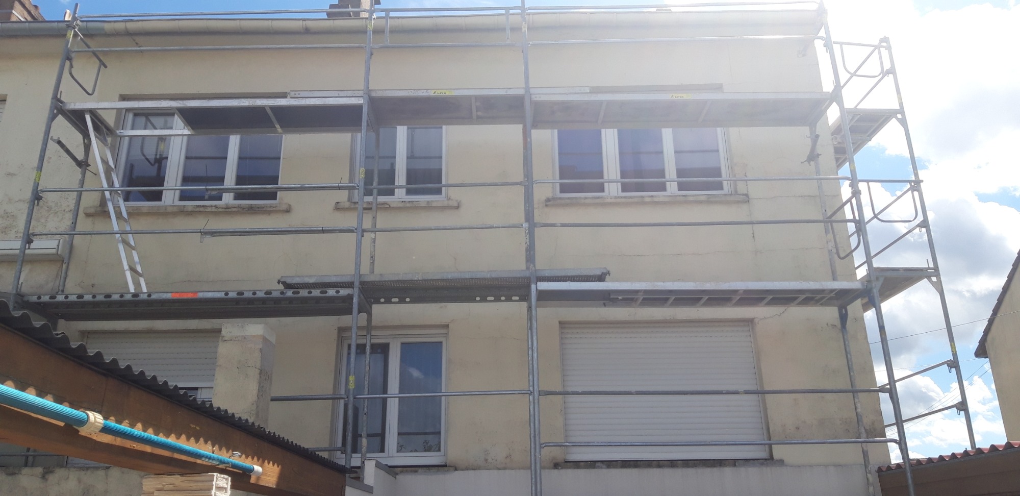 Dwupiętrowy dom w trakcie remontu elewacji, otoczony rusztowaniem, widoczne ślady wcześniejszych prac i przygotowania do kolejnych etapów.