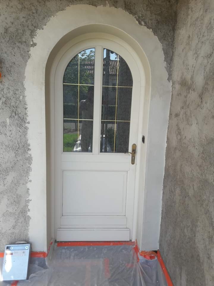 Białe, łukowe drzwi wejściowe z szybami i złotymi szprosami, osadzone w surowej elewacji w trakcie prac wykończeniowych, zabezpieczone folią malarską i taśmą.