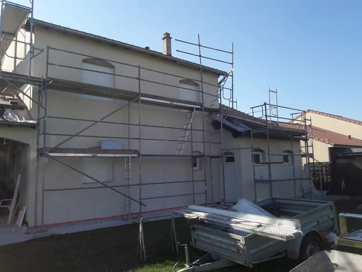 Dom jednorodzinny w trakcie tynkowania elewacji, widoczne rusztowania, przyczepka z materiałami budowlanymi i niezabezpieczony fragment dachu.