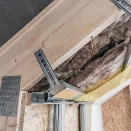CER&BUD Marcin Ceranka - Montaż wieszaków krokwiowych i izolacji z wełny mineralnej w konstrukcji dachu, widoczne elementy metalowe i drewniane, folia paroizolacyjna.