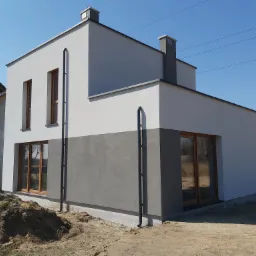 Dwukondygnacyjny budynek w Radostowicach z dwukolorową elewacją, jasnoszarą na górze i ciemnoszarą na dole, z drewnianymi ramami okiennymi i dwoma kominami na dachu, w otoczeniu ziemi i innych...