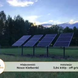 Instalacja fotowoltaiczna off-grid o mocy 3,84 kWp w Nowych Kiełbonkach, cztery panele na metalowych słupkach, w tle las i zachmurzone niebo o zmierzchu, widoczny fragment ogrodzenia i samochód.