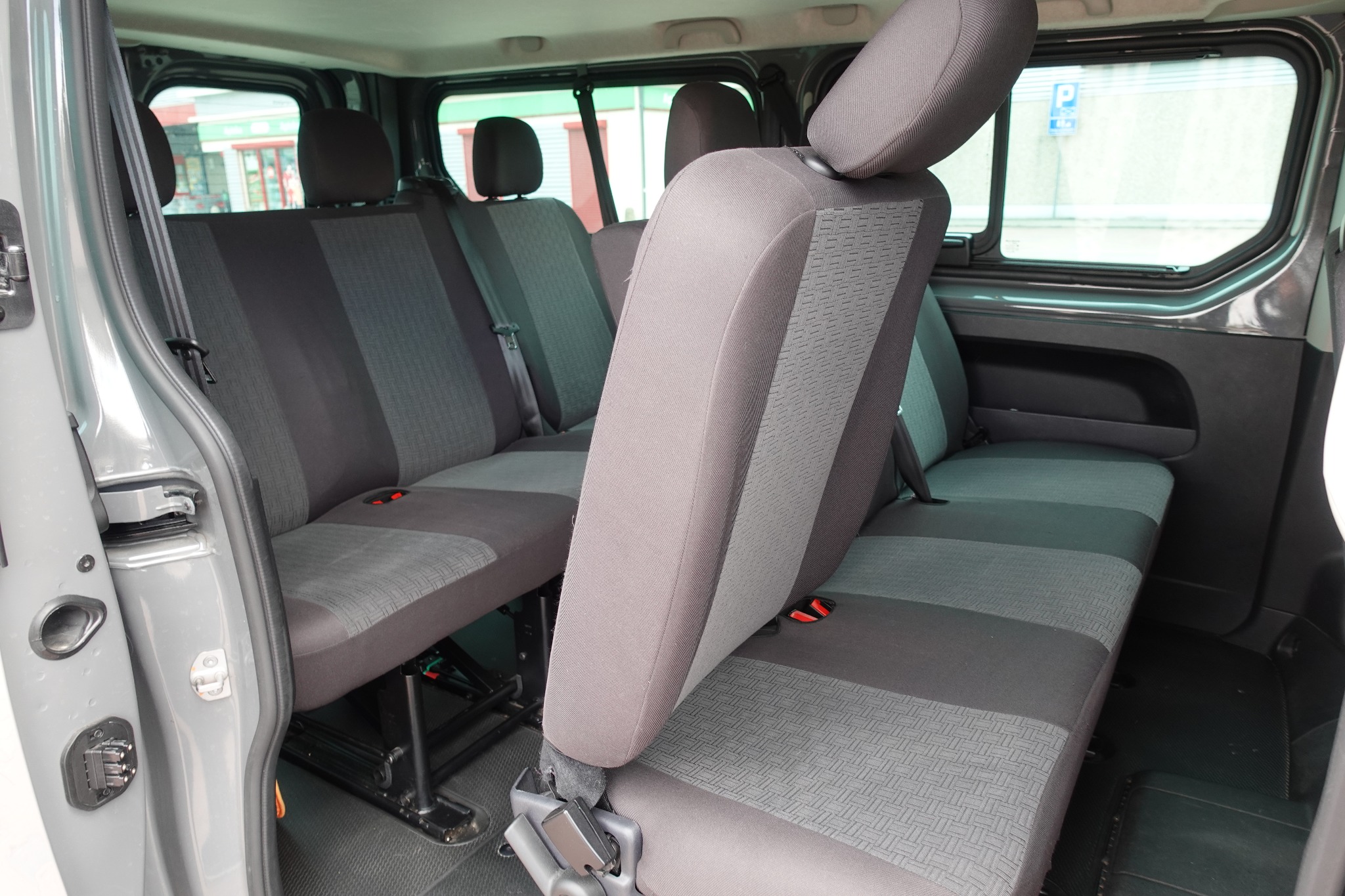 Wnętrze szarego busa z trzema rzędami siedzeń z szaro-czarną tapicerką, widok z otwartych drzwi bocznych na przestrzeń pasażerską.