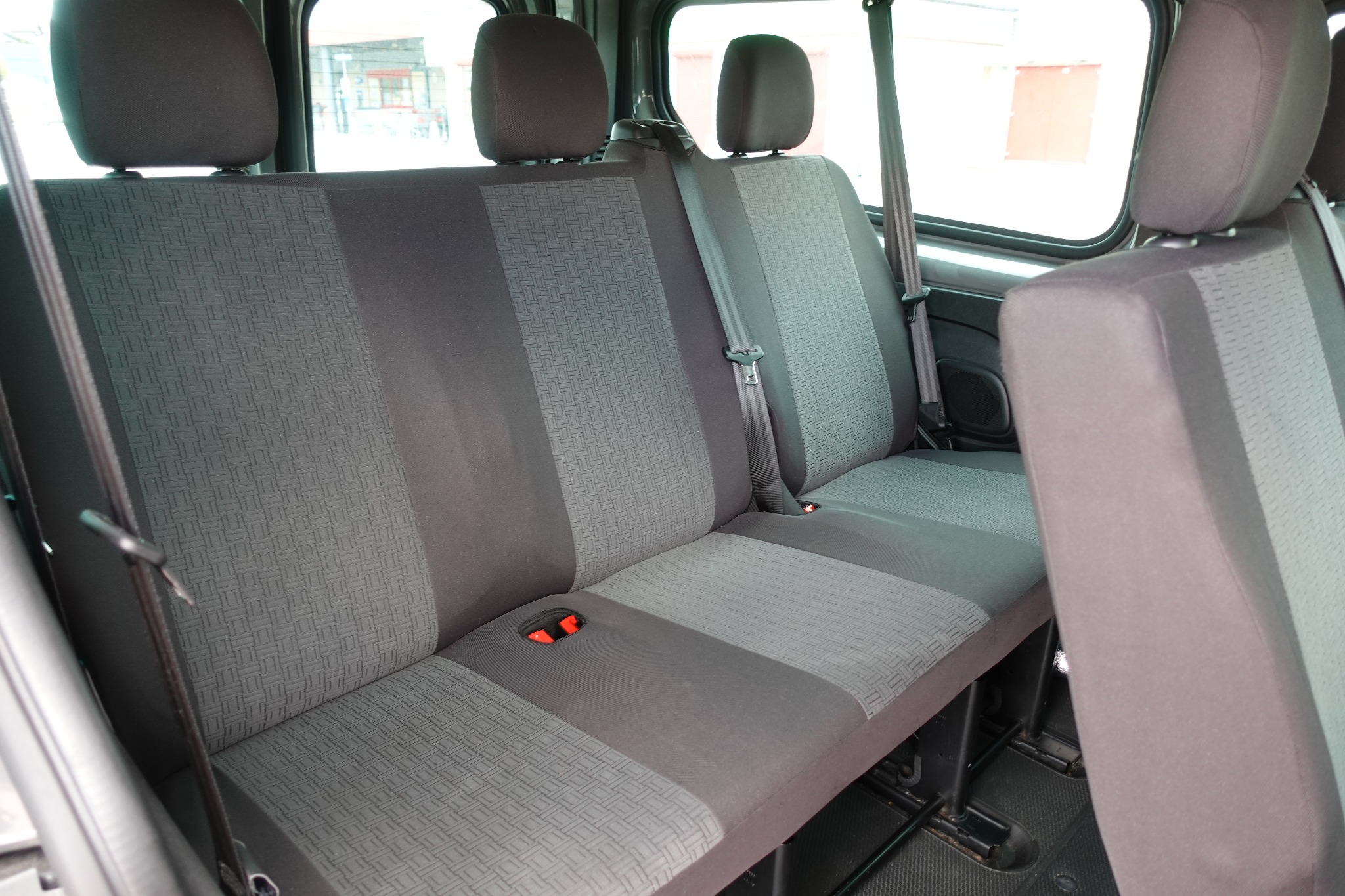 Wnętrze busa: trzyosobowa tylna kanapa z szarą tapicerką w geometryczny wzór, widoczne pasy bezpieczeństwa i uchwyty na napoje.