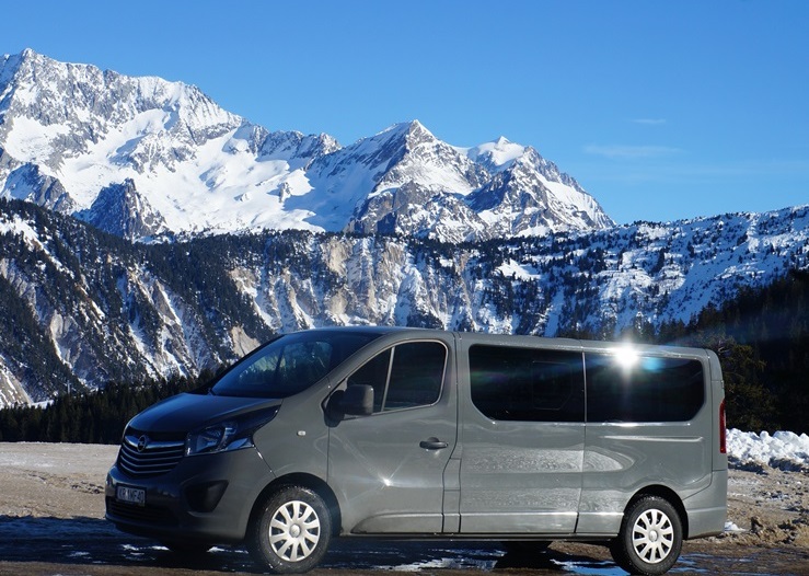 Szary van marki Opel Vivaro na tle ośnieżonych gór w słoneczny dzień, widoczny numer rejestracyjny KR 1MF40.
