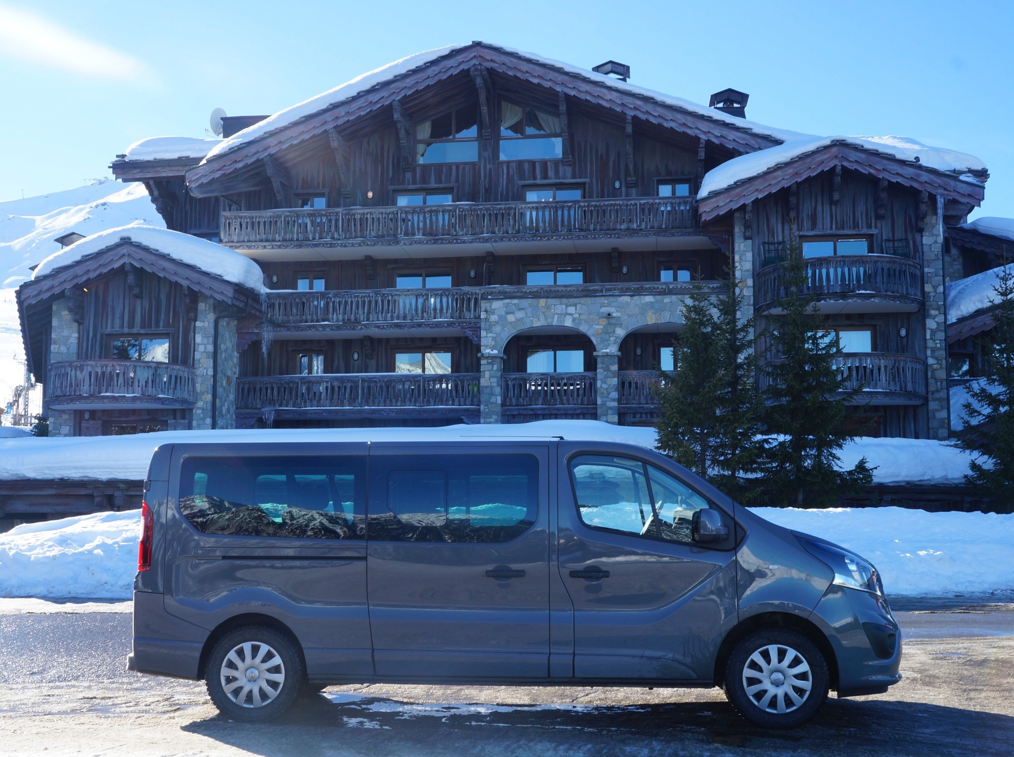 Szary van na tle drewnianego, wielopiętrowego budynku w zimowej scenerii górskiej, z odbiciem gór w szybach.