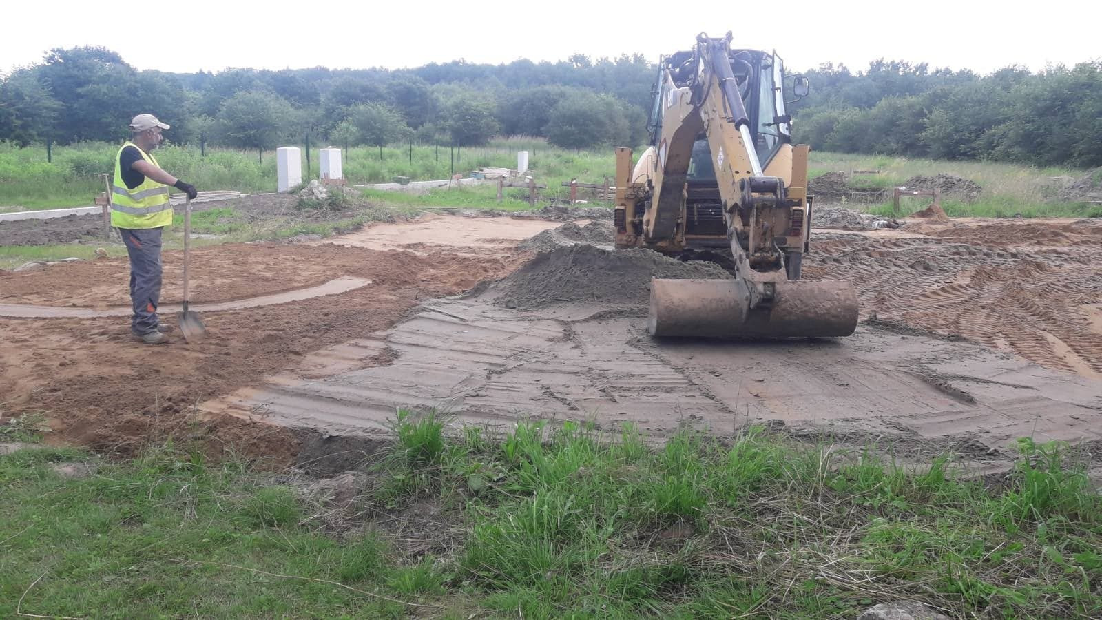 Prace ziemne: koparka wyrównuje teren pod fundamenty, pracownik z łopatą w odblaskowej kamizelce nadzoruje prace, widoczne białe słupki wyznaczające obrys budynku.