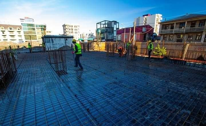 Zbrojenie płyty fundamentowej na budowie domu, widoczni pracownicy w kamizelkach odblaskowych, w tle budynki miejskie pod błękitnym niebem.