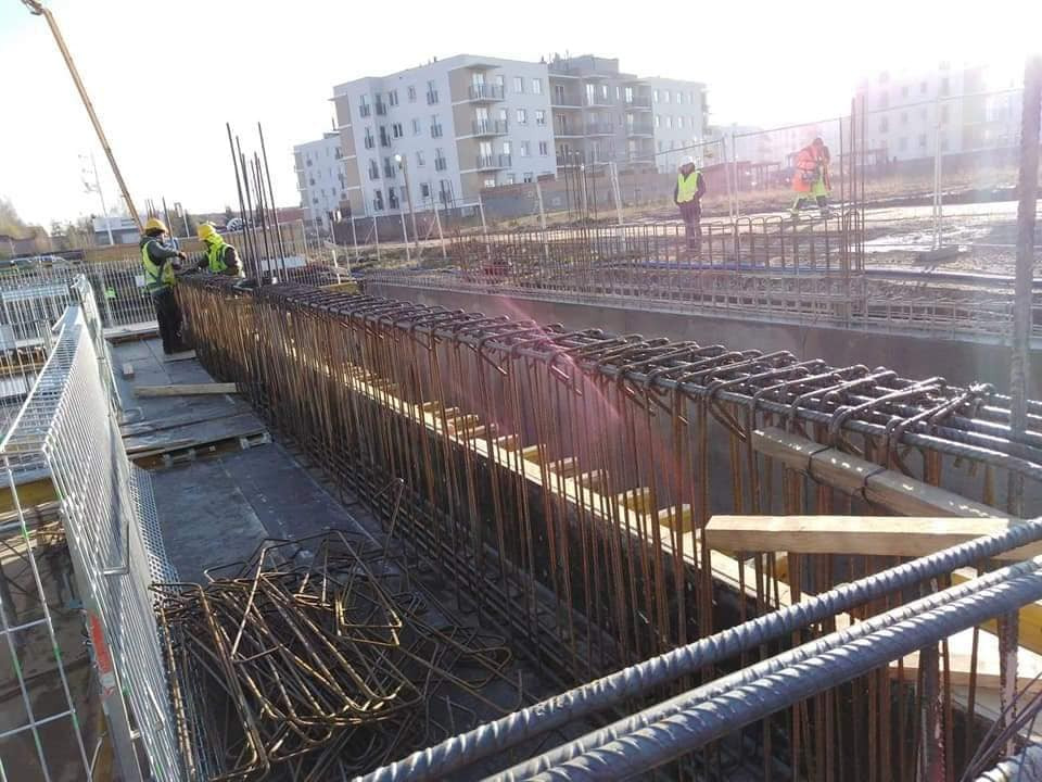 Zbrojenie fundamentów pod budynek wielorodzinny, widoczni pracownicy w żółtych kamizelkach i kaskach, w tle budynek mieszkalny i dźwig.