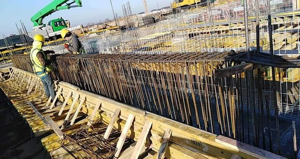 Zbrojenie w szalunku przygotowane do zalania betonem na budowie domu murowanego, widoczni pracownicy w kaskach i kamizelkach odblaskowych, w tle maszyny budowlane.