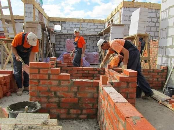 Murarze w trakcie wznoszenia ścian zewnętrznych domu z cegły klinkierowej na placu budowy, widoczne bloczki betonowe i drewniane rusztowania.