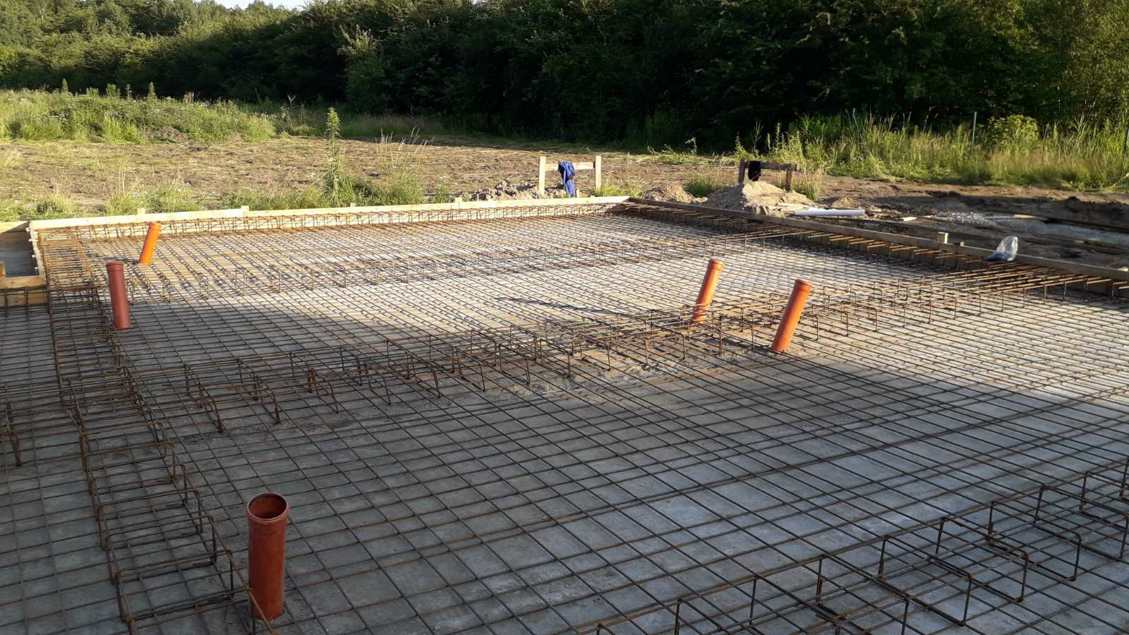 Zbrojenie fundamentu w formie siatki z wystającymi pomarańczowymi rurami, drewniany szalunek obwodowy, w tle teren budowy i zieleń.