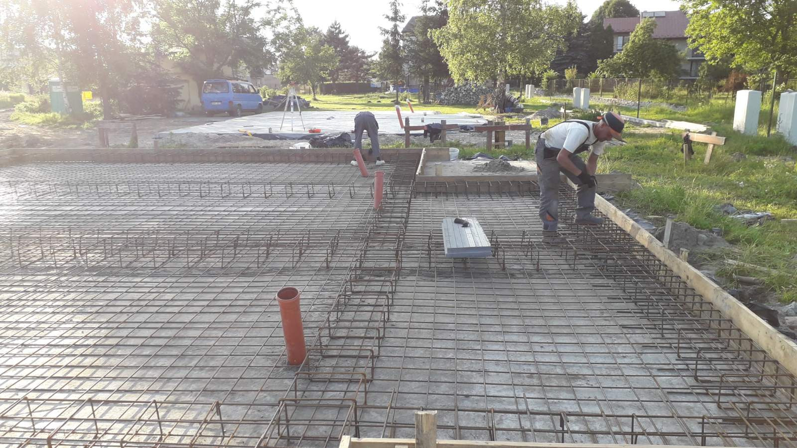 Zbrojenie fundamentów domu jednorodzinnego z wystającymi rurami kanalizacyjnymi, widoczni dwaj pracownicy w trakcie montażu, teren budowy z zielenią w tle.