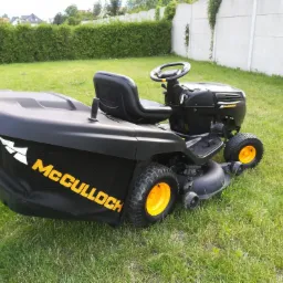 Czarna kosiarka traktorowa McCulloch z żółtymi kołami, stojąca na trawniku przed żywopłotem i betonowym murem.