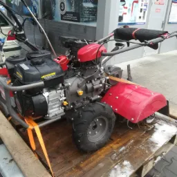 Glebogryzarka spalinowa z czerwonymi elementami, zabezpieczona pomarańczowymi pasami transportowymi na drewnianej palecie przyczepki samochodowej, tło stanowi myjnia samochodowa.