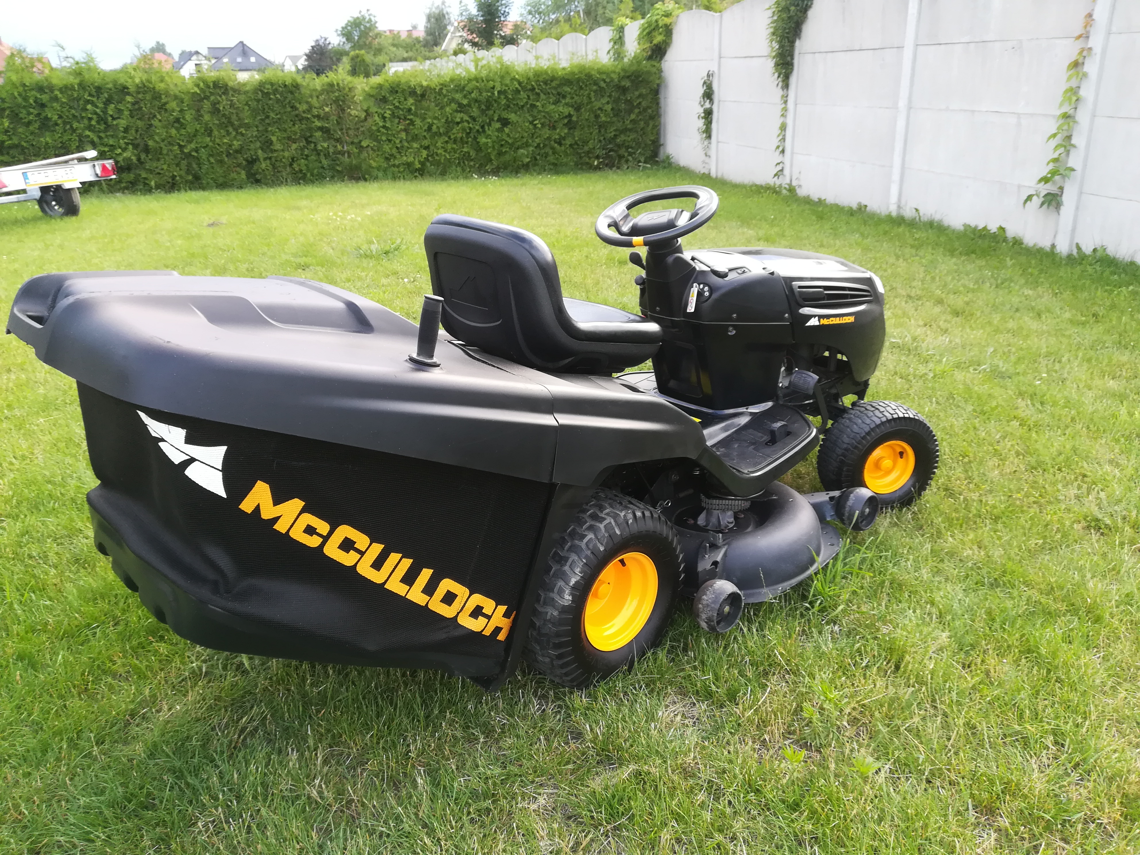 Czarna kosiarka traktorowa McCulloch z żółtymi kołami, stojąca na trawniku przed żywopłotem i betonowym murem.
