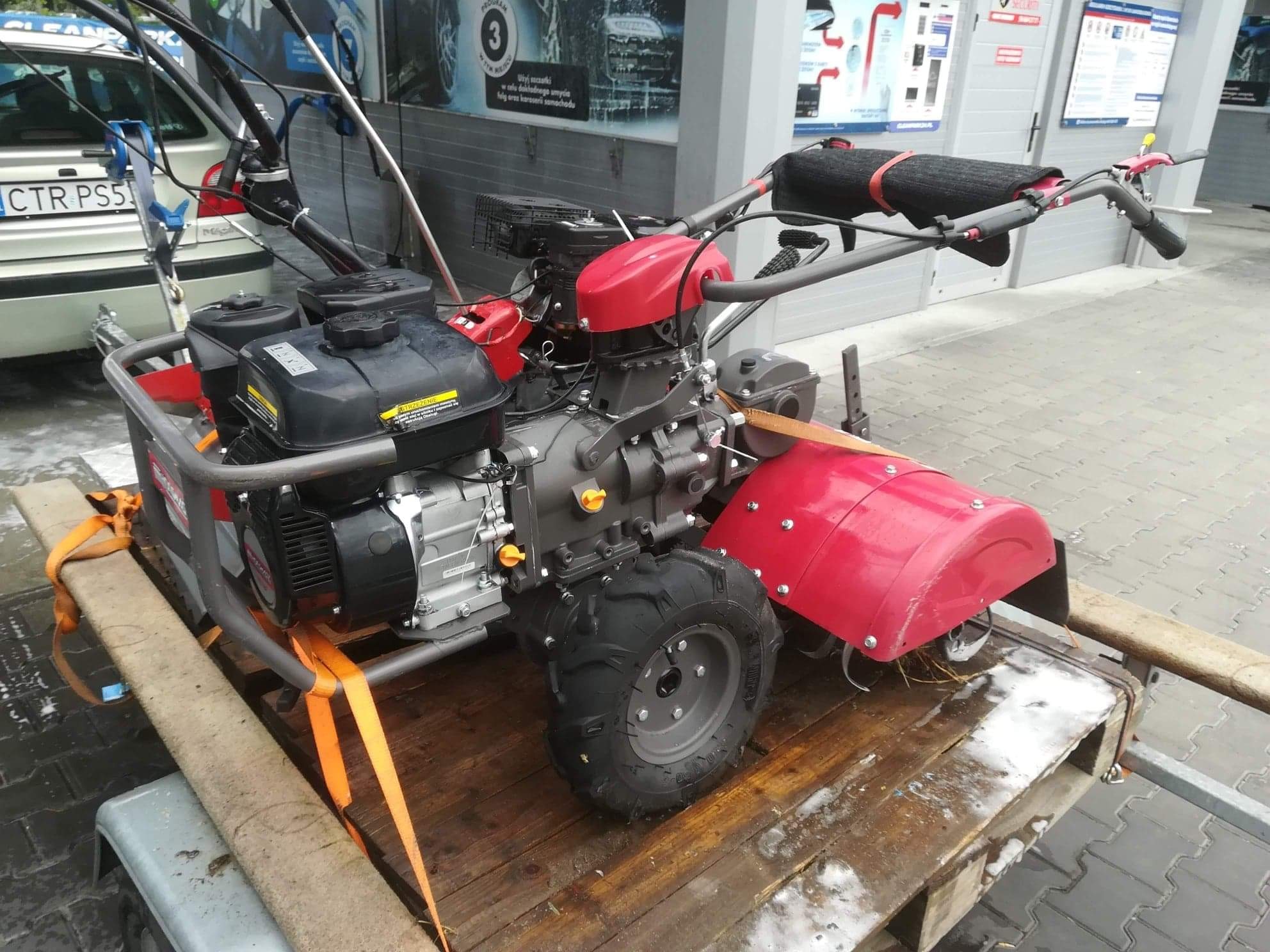 Glebogryzarka spalinowa z czerwonymi elementami, zabezpieczona pomarańczowymi pasami transportowymi na drewnianej palecie przyczepki samochodowej, tło stanowi myjnia samochodowa.