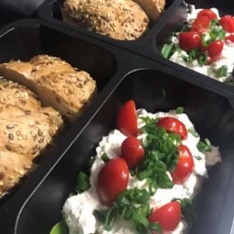 Porcje w czarnych pojemnikach: pieczywo pełnoziarniste i pasta twarogowa z pomidorkami koktajlowymi i szczypiorkiem na sałacie.