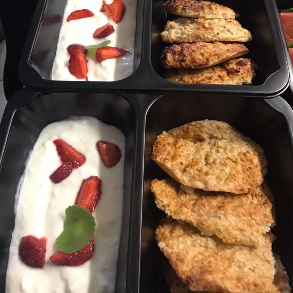 Dwa czarne pojemniki z jedzeniem: jogurt naturalny z kawałkami truskawek i listkiem mięty oraz pieczone kotlety owsiane.