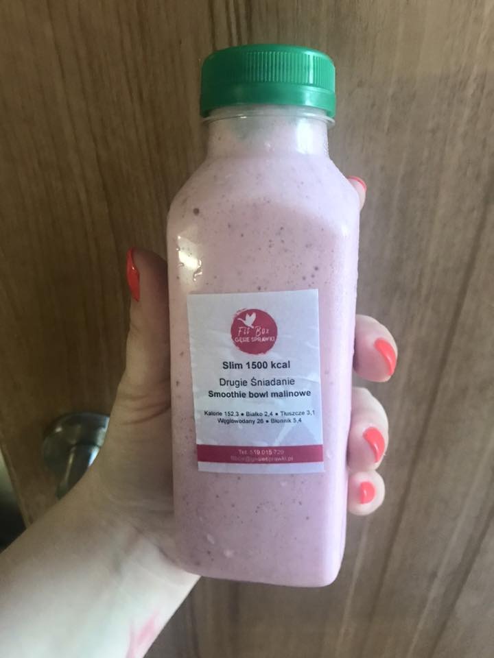 Różowy smoothie bowl malinowy w plastikowej butelce z zieloną zakrętką, trzymany w dłoni z pomalowanymi na czerwono paznokciami, na etykiecie informacje o kaloryczności i składnikach odżywczych.