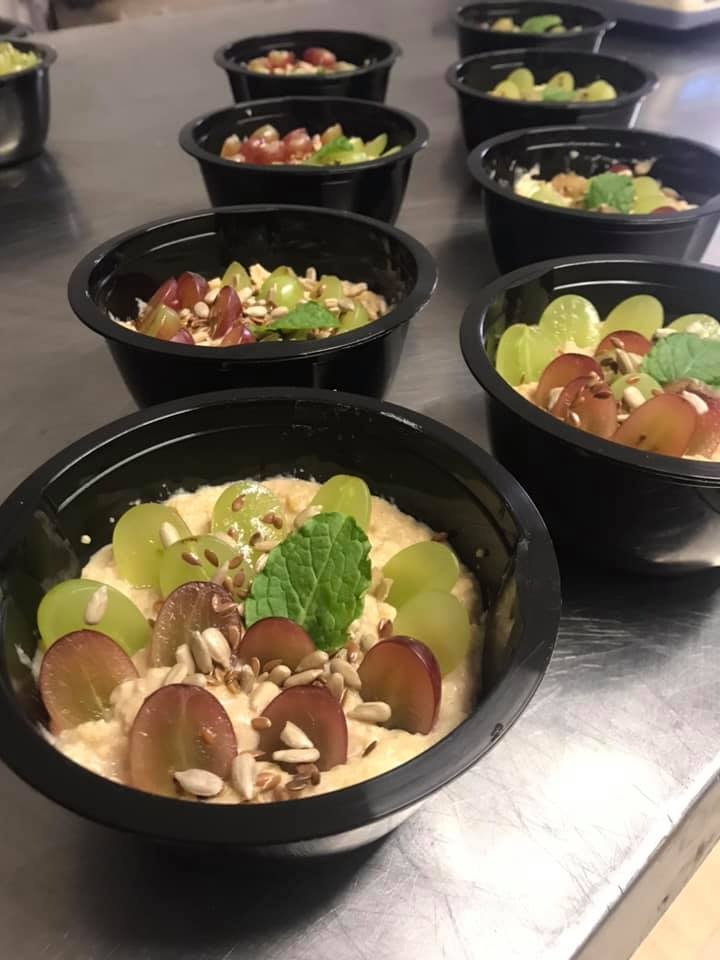 Porcje deseru w czarnych miseczkach z kremem, zielonymi i czerwonymi winogronami, nasionami słonecznika i listkiem mięty, ułożone rzędami na metalowym blacie.